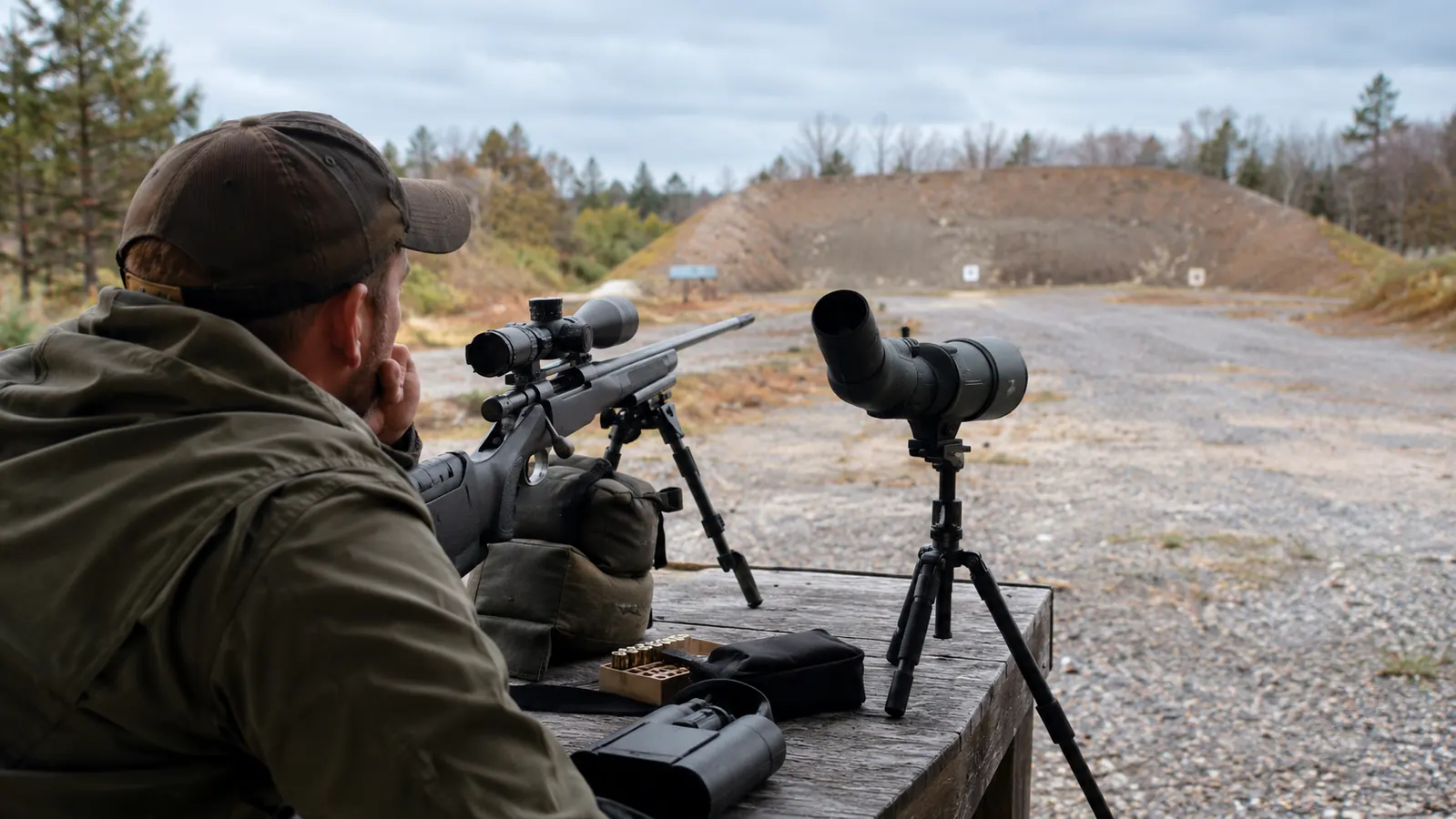 A hunter standing at a shooting bench with a rifle and a spotting scope, looking toward a 200-yard berm