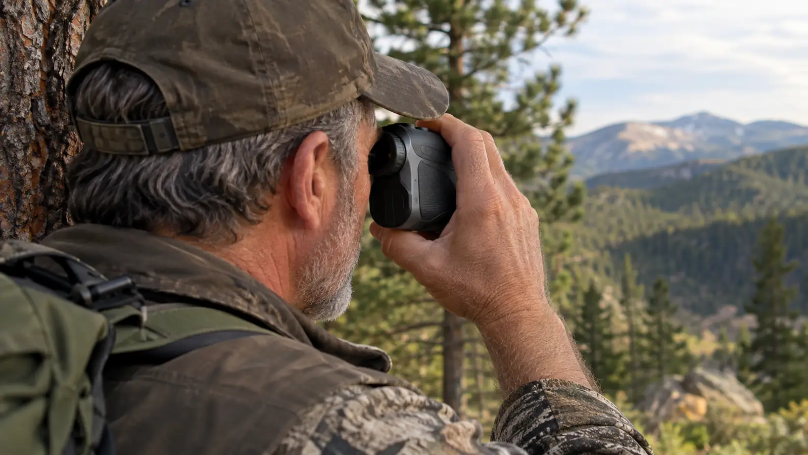 A hand holding a compact camo rangefinder up to a hunter's eye while he leans against a pine trunk