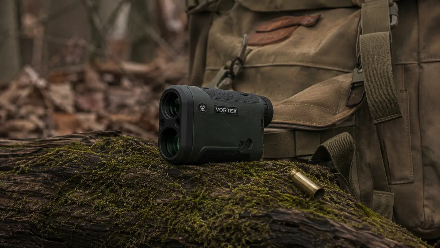 A rangefinder lying on a mossy log beside a worn hunting pack and a spent cartridge