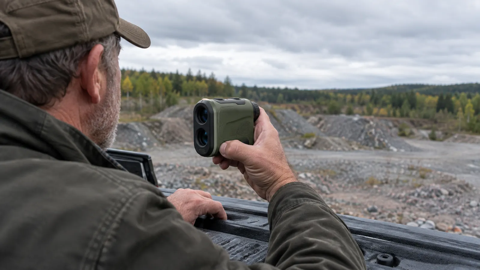 A hunter's hands holding a compact rangefinder steady against a truck tailgate, looking toward a far tree line