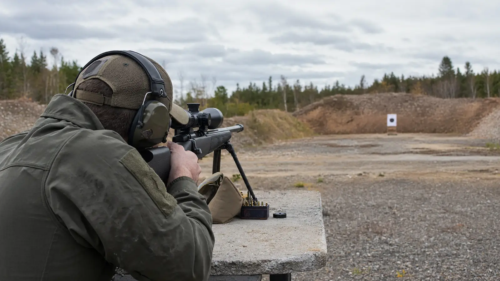 A hunter kneeling at a shooting bench with a bolt-action rifle, looking through a riflescope at a 100-yard berm
