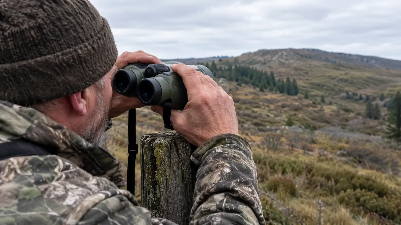 A hunter's hands holding binoculars steady against a fence post while glassing a distant hillside
